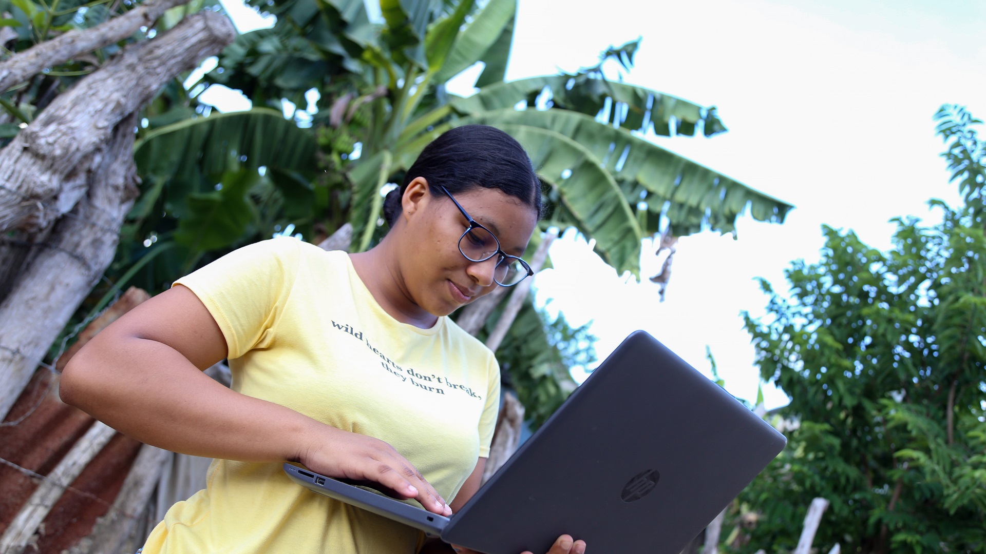 fotografía: Una joven en zona rural, usa el computador portátil de pie. Con su mano izquierda lo sostiene y con la derecha, digita en el teclado.